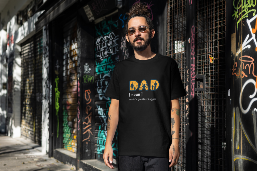 Man wearing an oversized black t-shirt with the text "Dad [noun] World's Greatest Hugger" and a silhouette design of a dad with kids, by Attire Dot